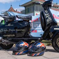 Black scooter with 'John Player Special' branding, two Union Jack helmets, and a blurred background with people and vehicles.