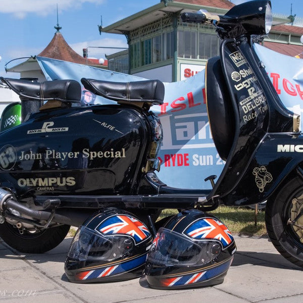 Black scooter with 'John Player Special' branding, two Union Jack helmets, and a blurred background with people and vehicles.